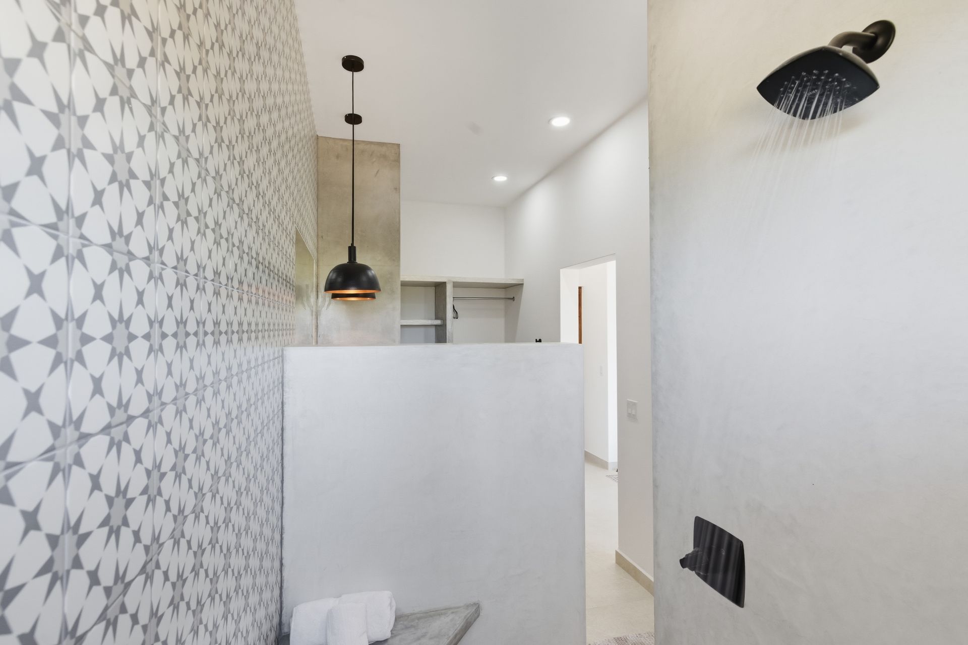 A modern, minimalist bathroom with patterned gray tile on one wall, a concrete shower partition, a matte black showerhead.