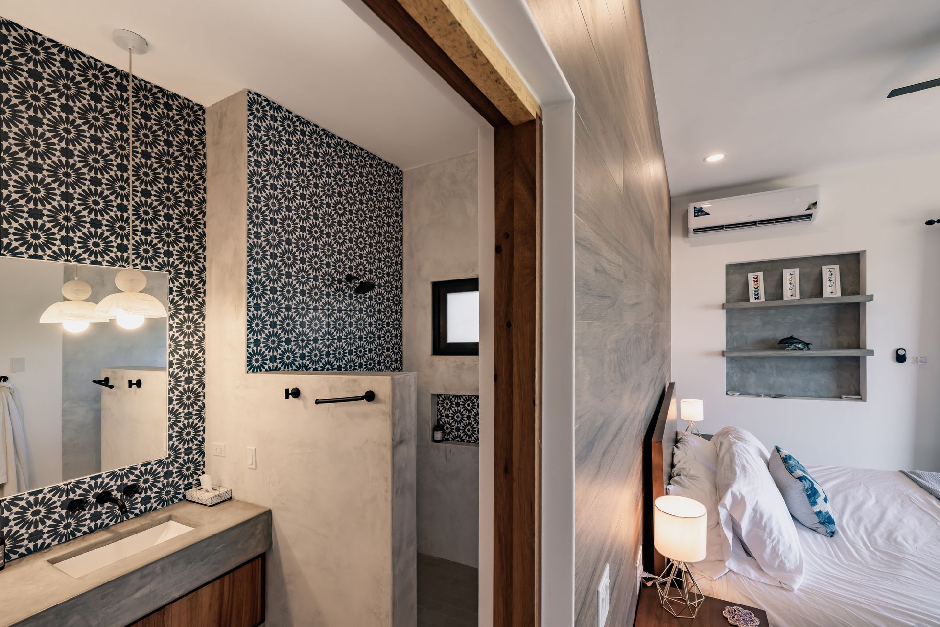 A view from a bedroom into an adjacent bathroom with blue patterned tile walls, a concrete vanity, and a wooden bed frame.