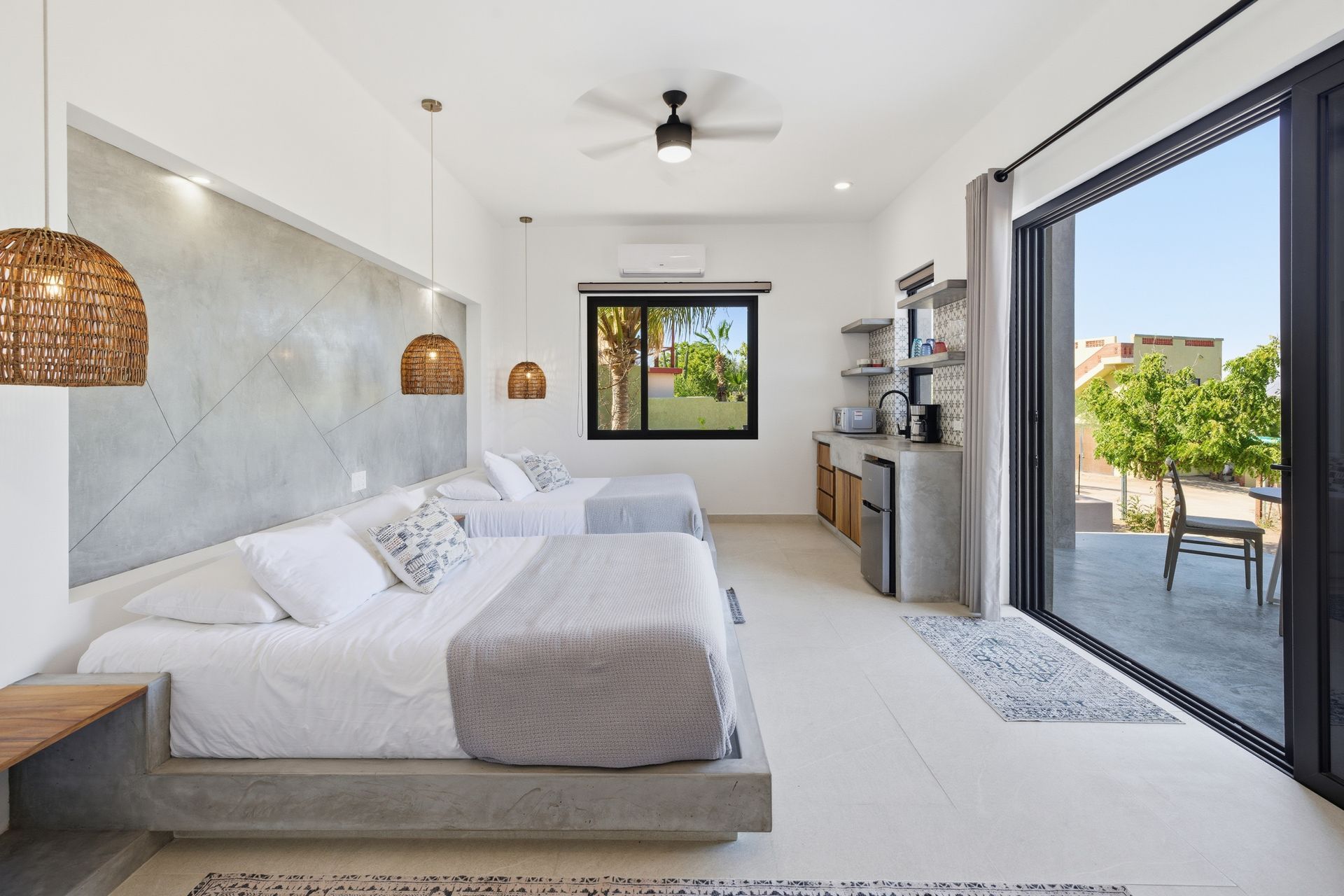 A bright, modern studio room with two queen beds, minimalist gray concrete decor, kitchenette, and a large balcony door.