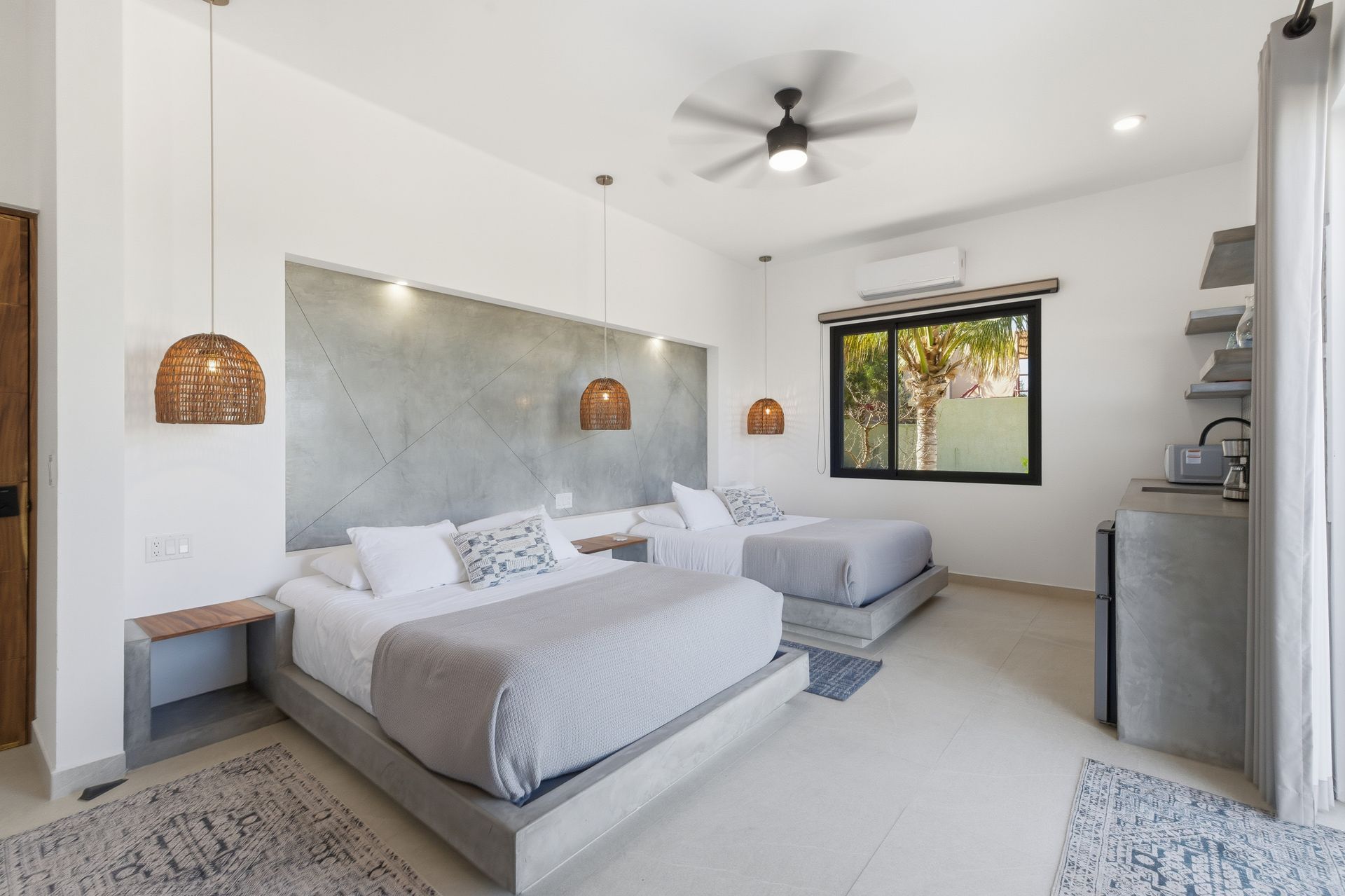 Modern bedroom with two beds, gray concrete feature wall, woven pendant lights, and a window overlooking greenery.