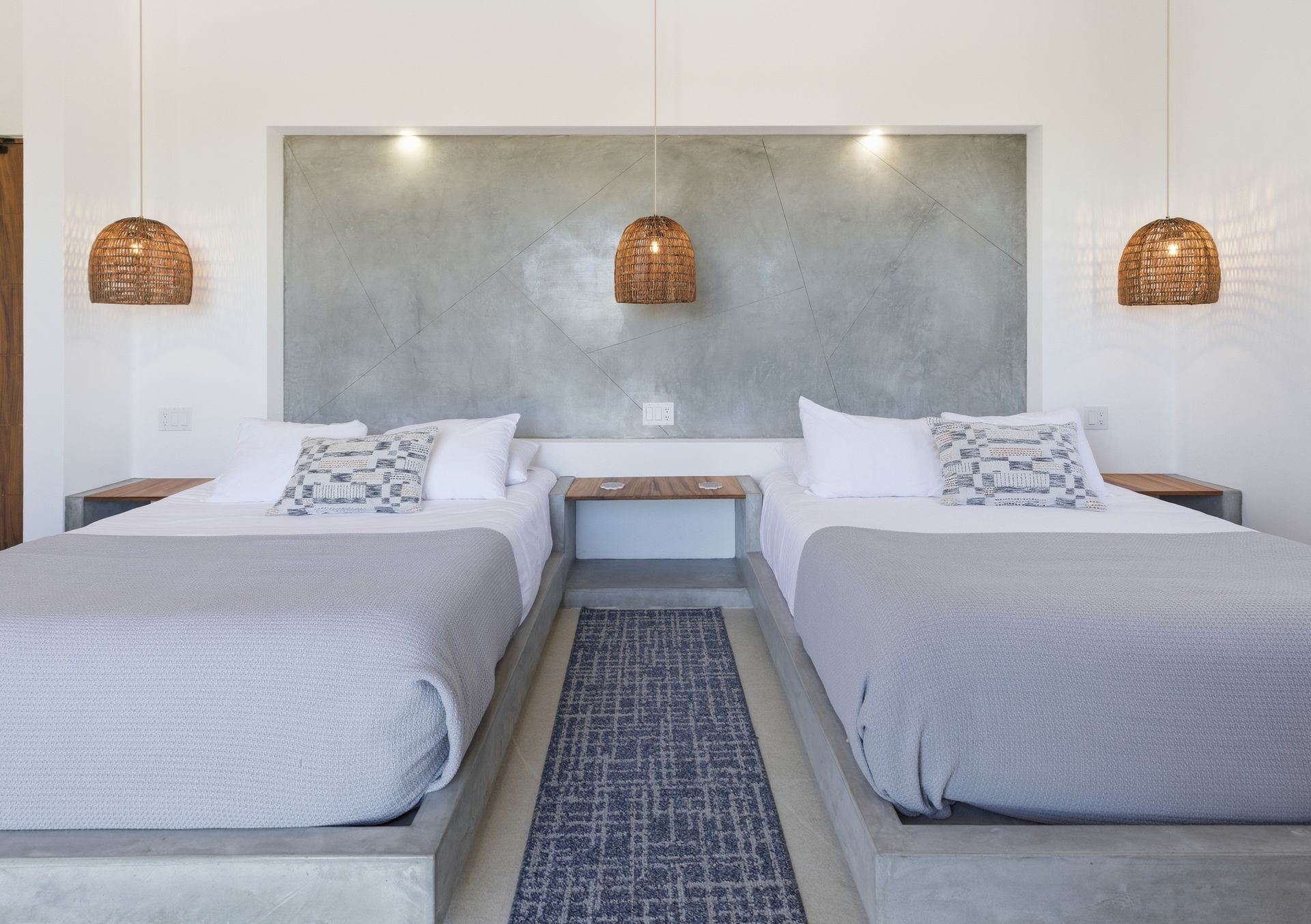 A hotel bedroom featuring two beds with gray bedding, centered by a concrete headboard wall and three hanging wicker lamps.