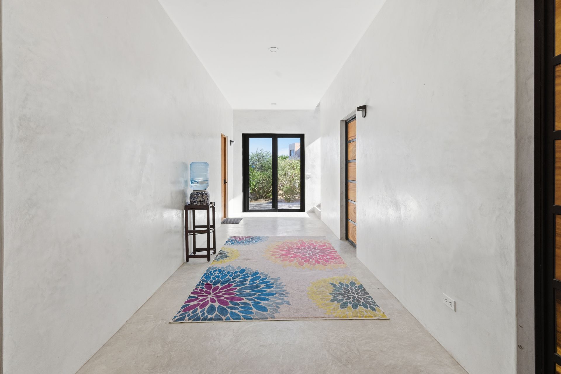 A long, white-walled hallway with polished concrete floors, a colorful floral rug, and a water cooler near a door.