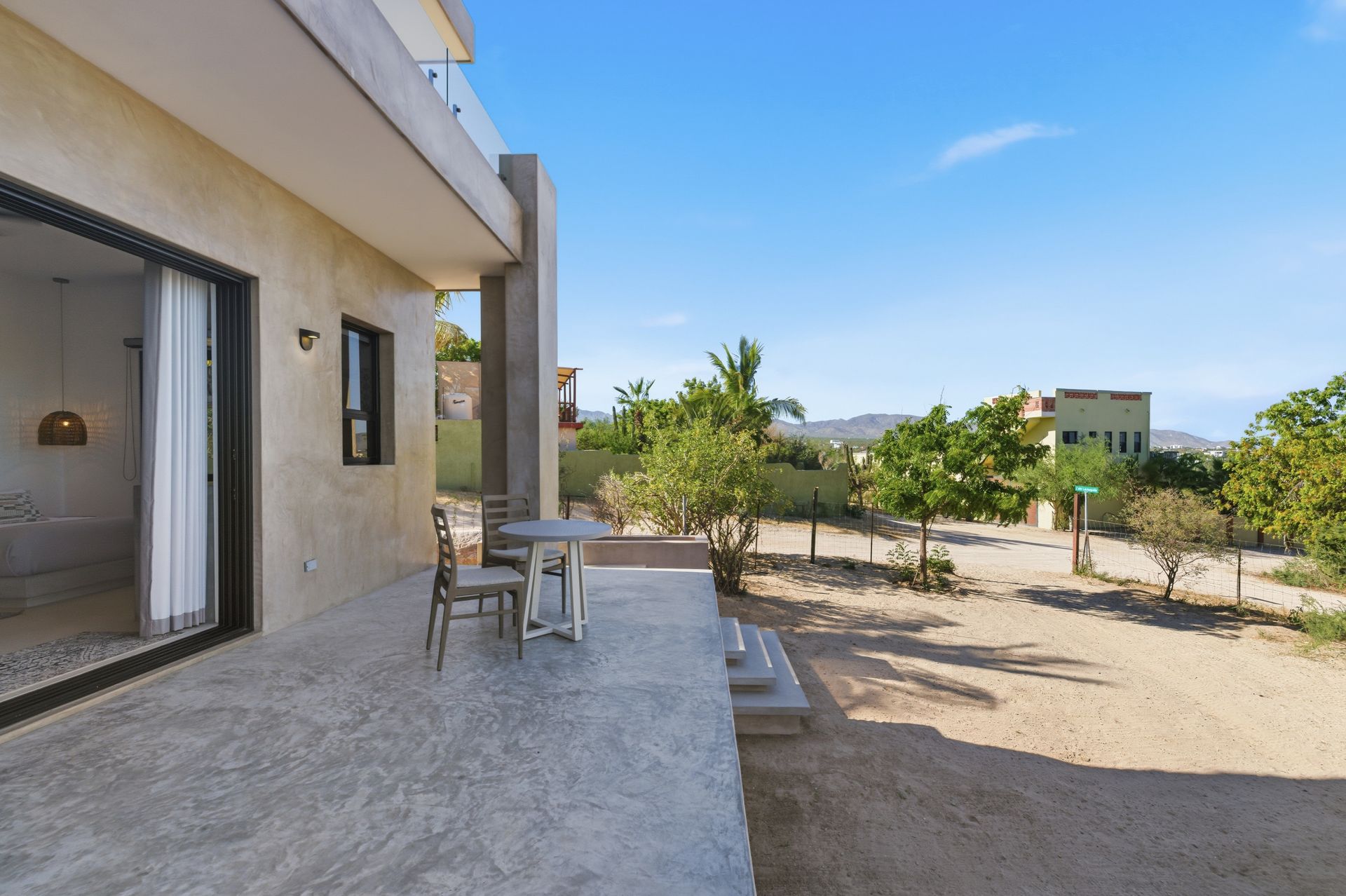 A modern patio with a small table and chairs extends from a house into a sunny, dry landscape with sparse green trees.