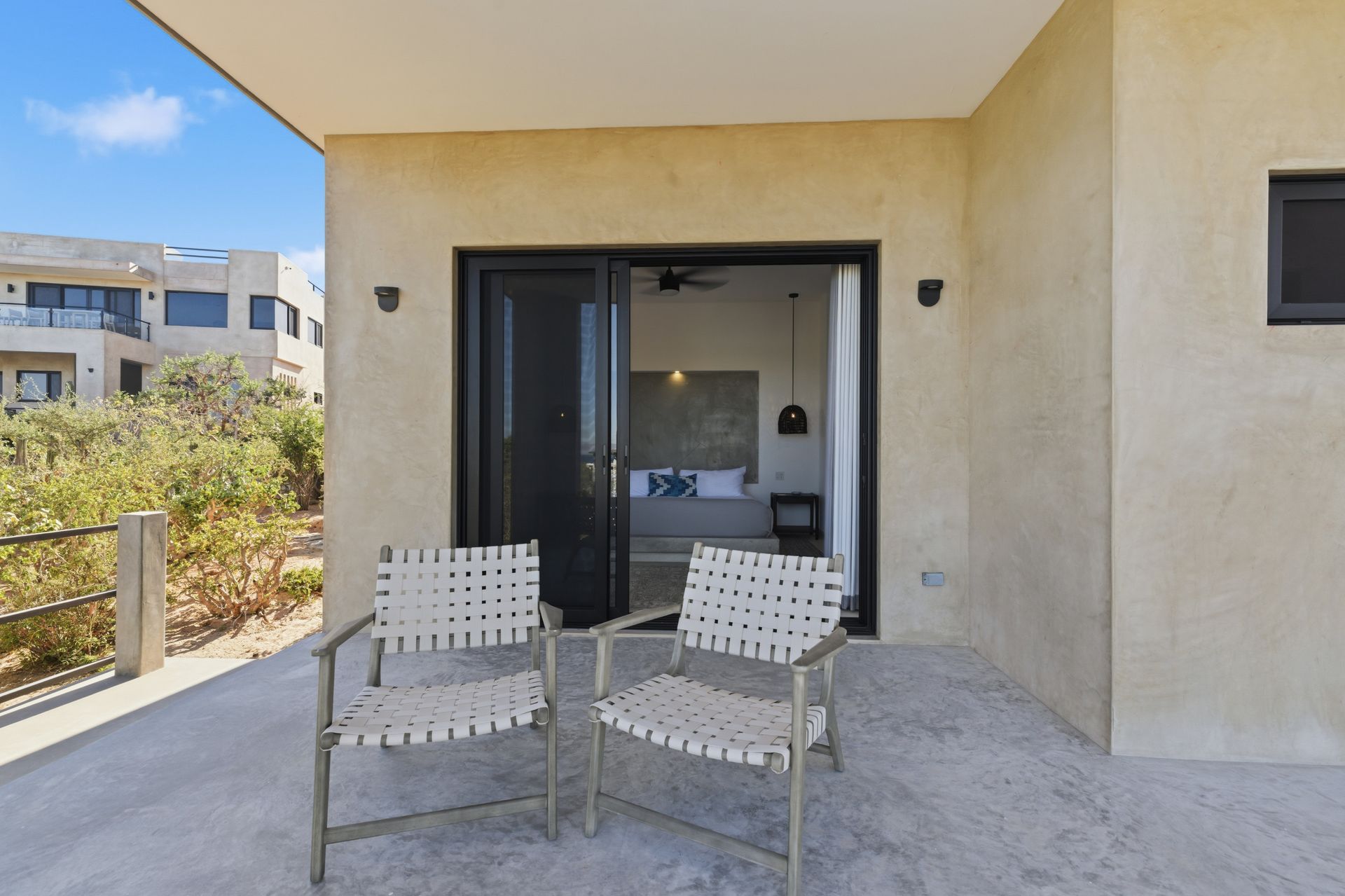 Two woven chairs on a concrete patio leading into a bedroom with a view of nearby buildings and trees.