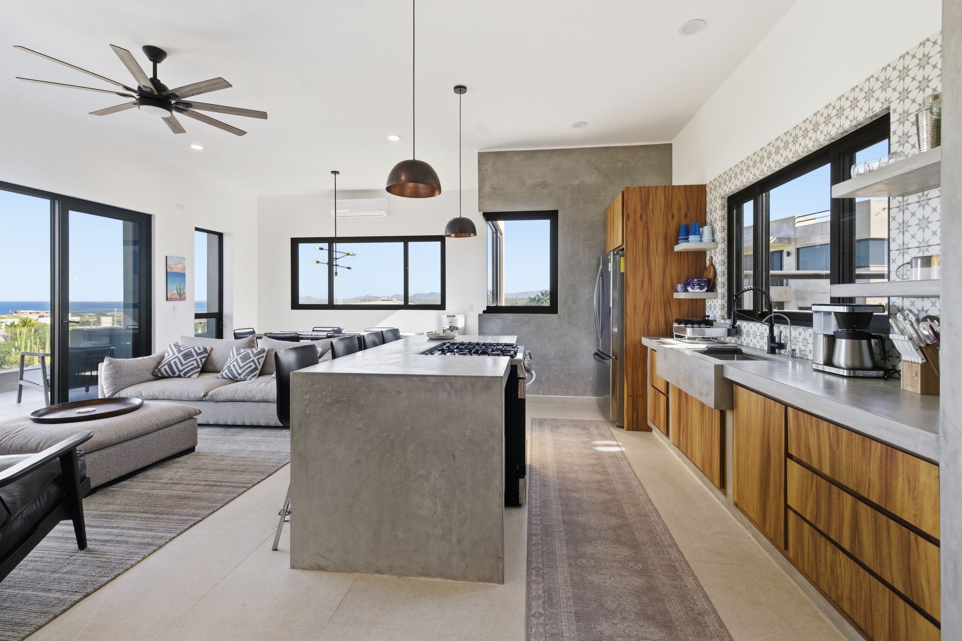 Open-concept modern kitchen and living area with a concrete island, wood cabinets, and large windows overlooking the ocean.