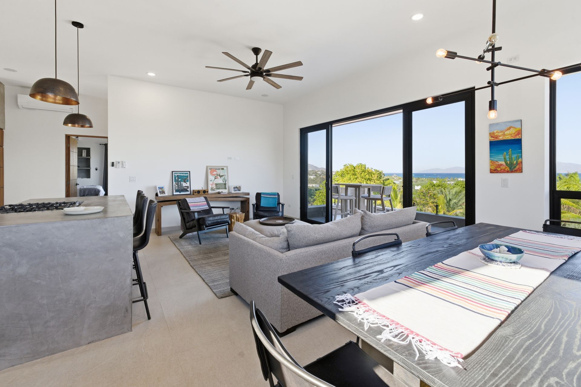 Modern open-plan living room with kitchen island, dining table, sofa, and floor-to-ceiling windows overlooking the ocean.