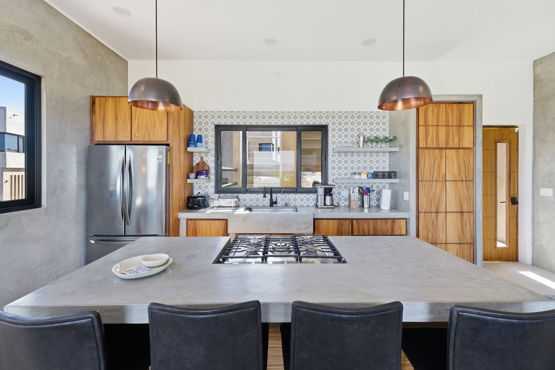 Modern kitchen with a gray concrete island and stovetop, wood cabinets, a stainless refrigerator, and pendant lights.