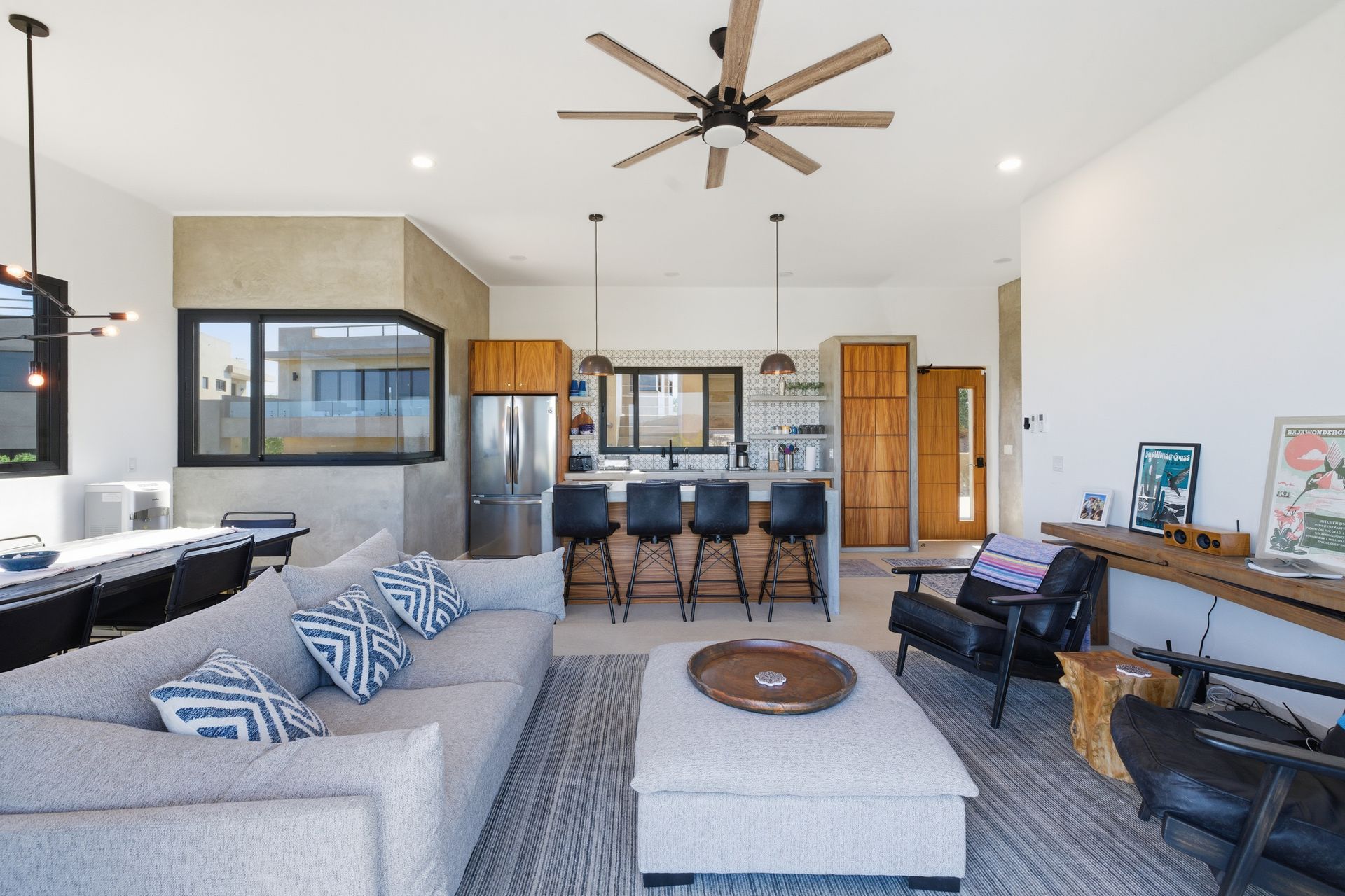 Open-concept living room with a grey sofa, ottoman, black chairs, kitchen island with stools, and wooden cabinetry.