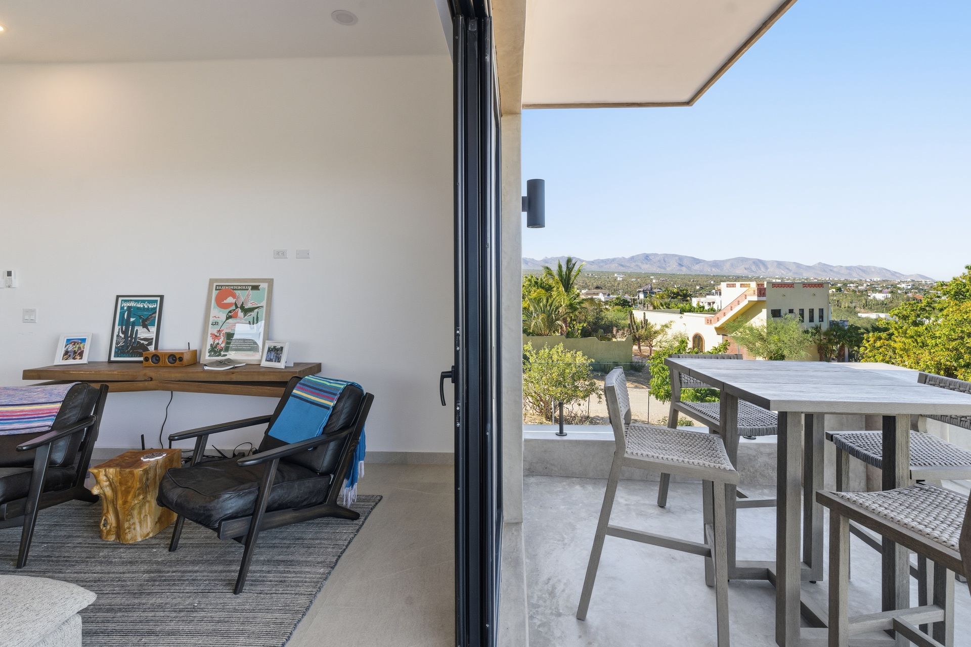 A living area flows into a bright, covered balcony with an outdoor dining set, overlooking hills under a clear blue sky.
