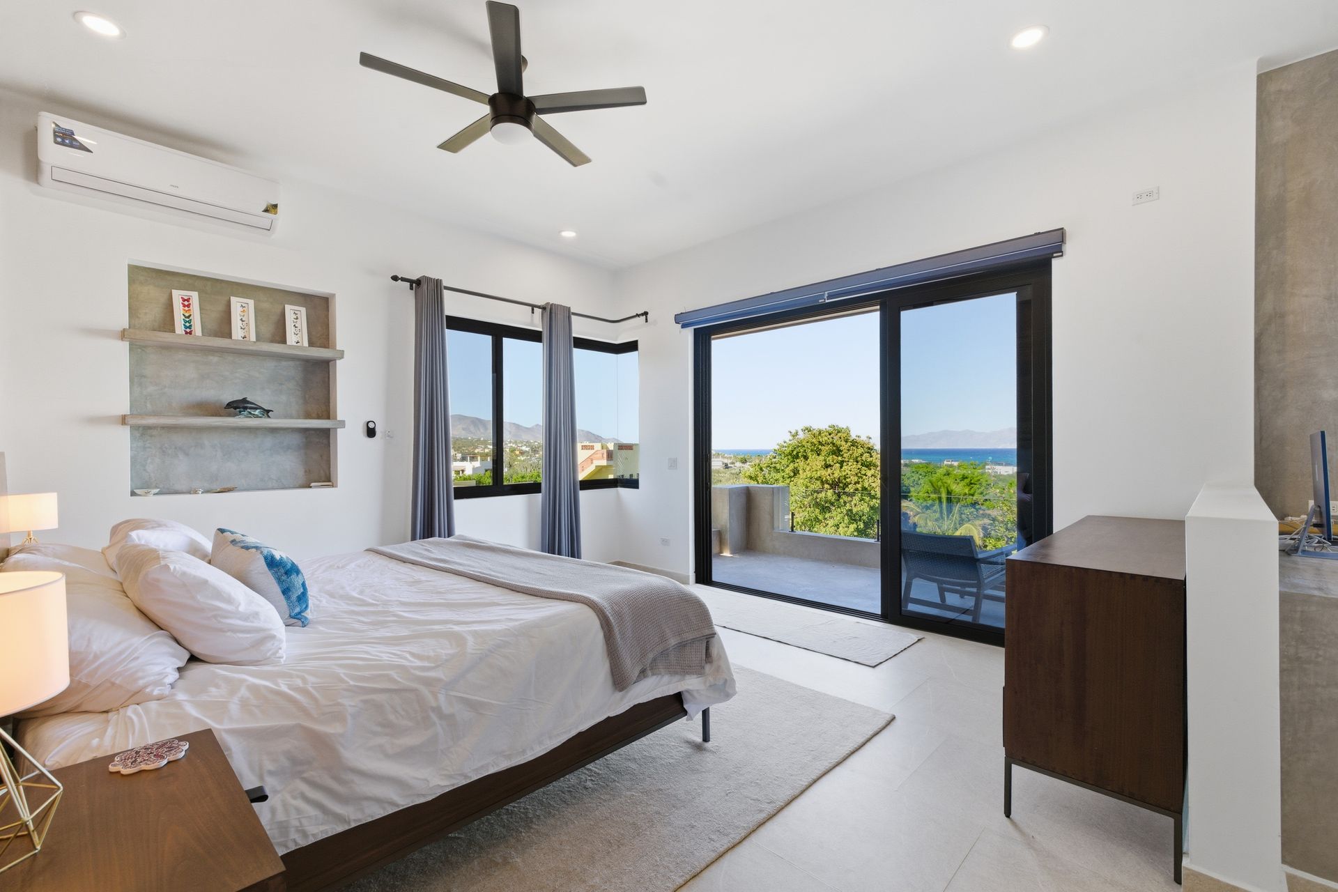 A modern, bright bedroom with a queen bed, shelving, ceiling fan, and sliding glass doors opening to an ocean view.
