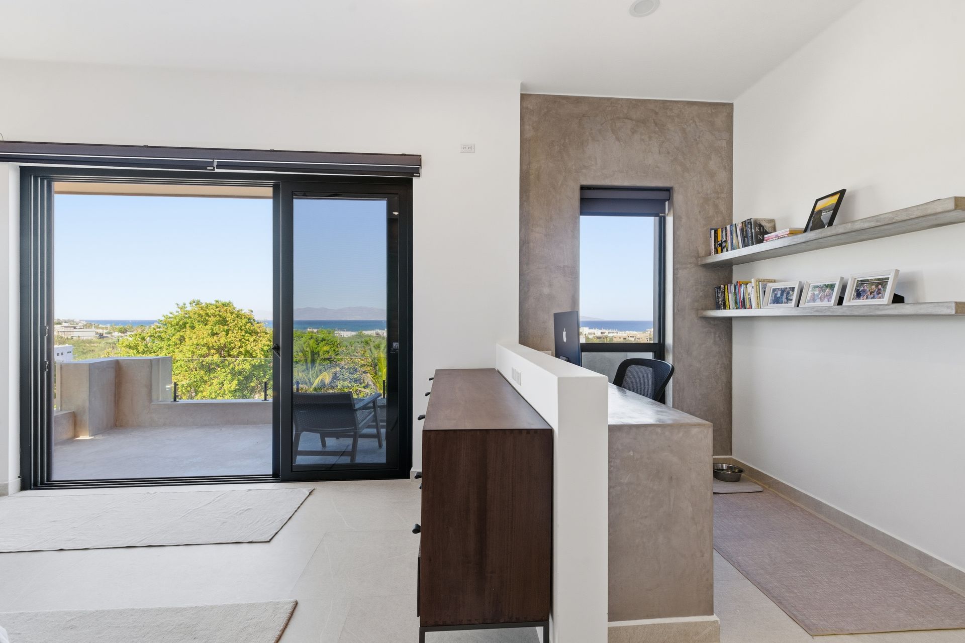 A minimalist home office with a modern desk, a wooden cabinet, shelves, and a large sliding door opening to a sea view.