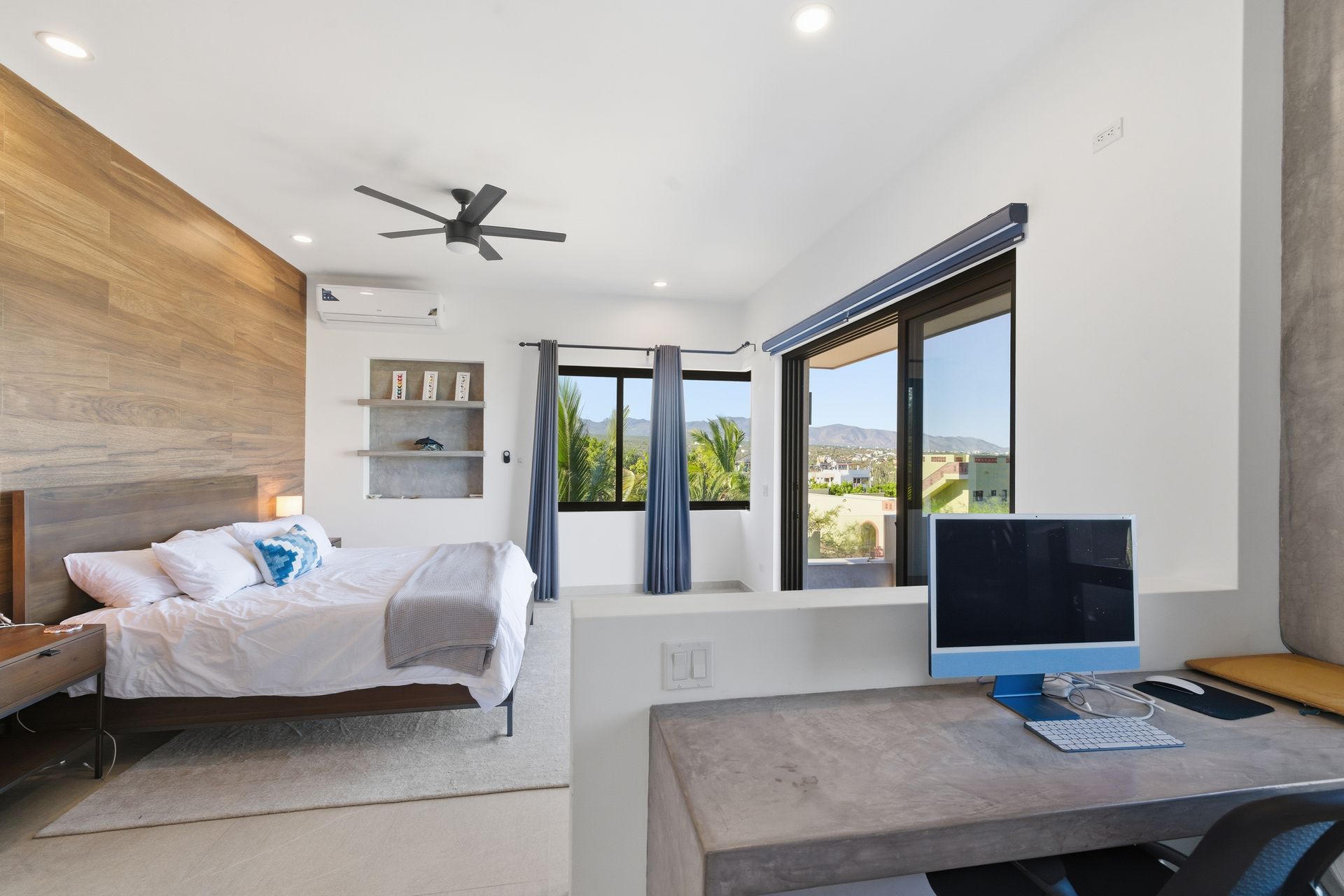 Modern bedroom with a wood accent wall, bed, desk with a monitor, and a large window overlooking a sunny outdoor area.