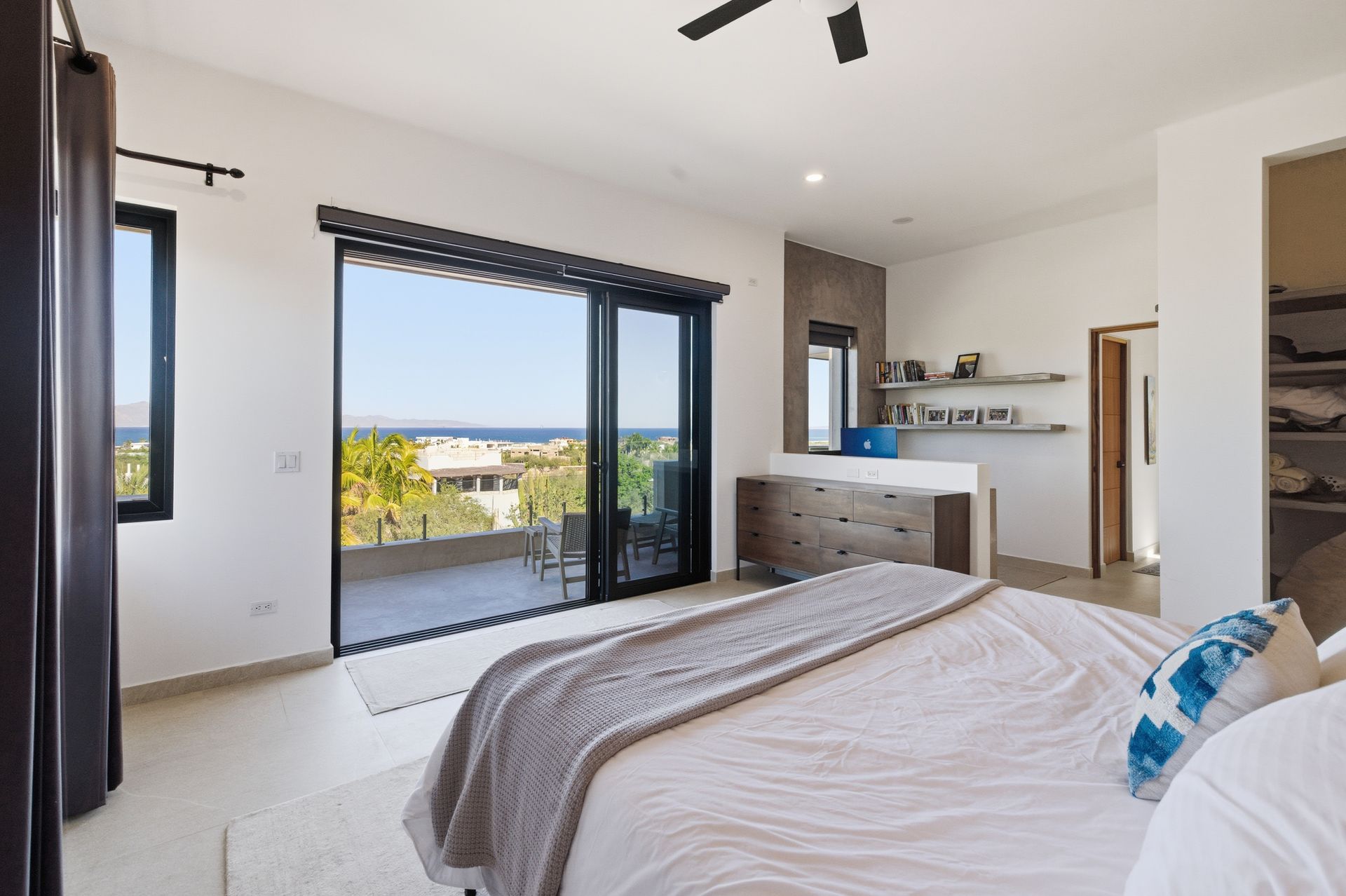 A bright, modern bedroom with a large bed, sliding glass doors opening to a scenic balcony, and an en-suite bathroom area.