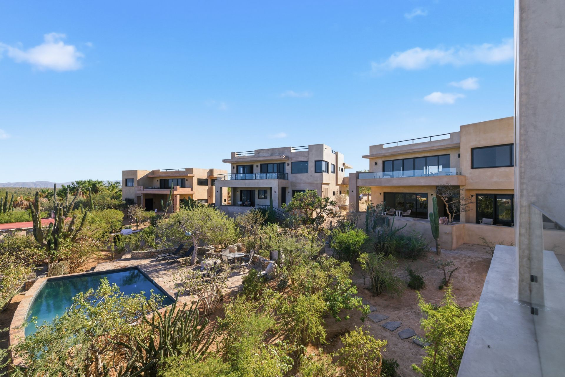 Modern desert villas with flat roofs and swimming pools surrounded by native scrubland under a clear blue sky.