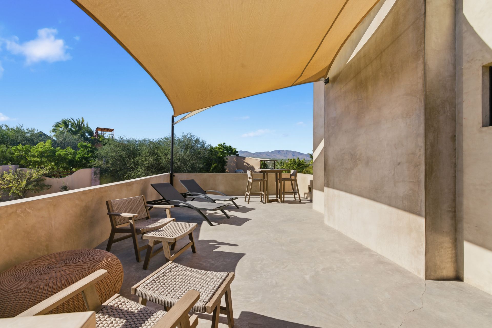 A sunlit outdoor terrace with patio furniture and a large beige shade sail against a clear blue sky.