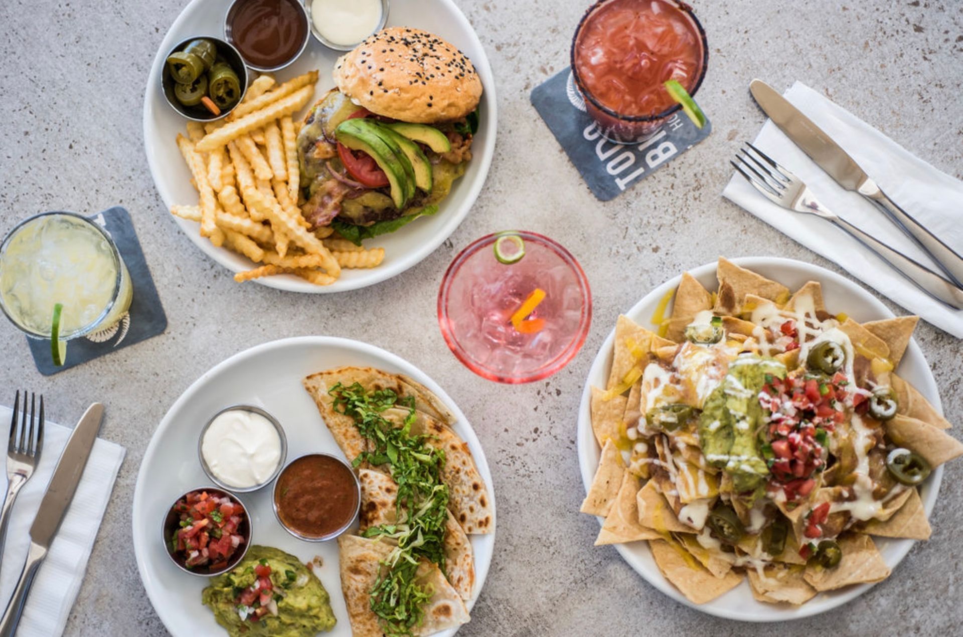 A top-down view of a spread featuring a burger with fries, a plate of quesadillas, nachos, and various colorful drinks.