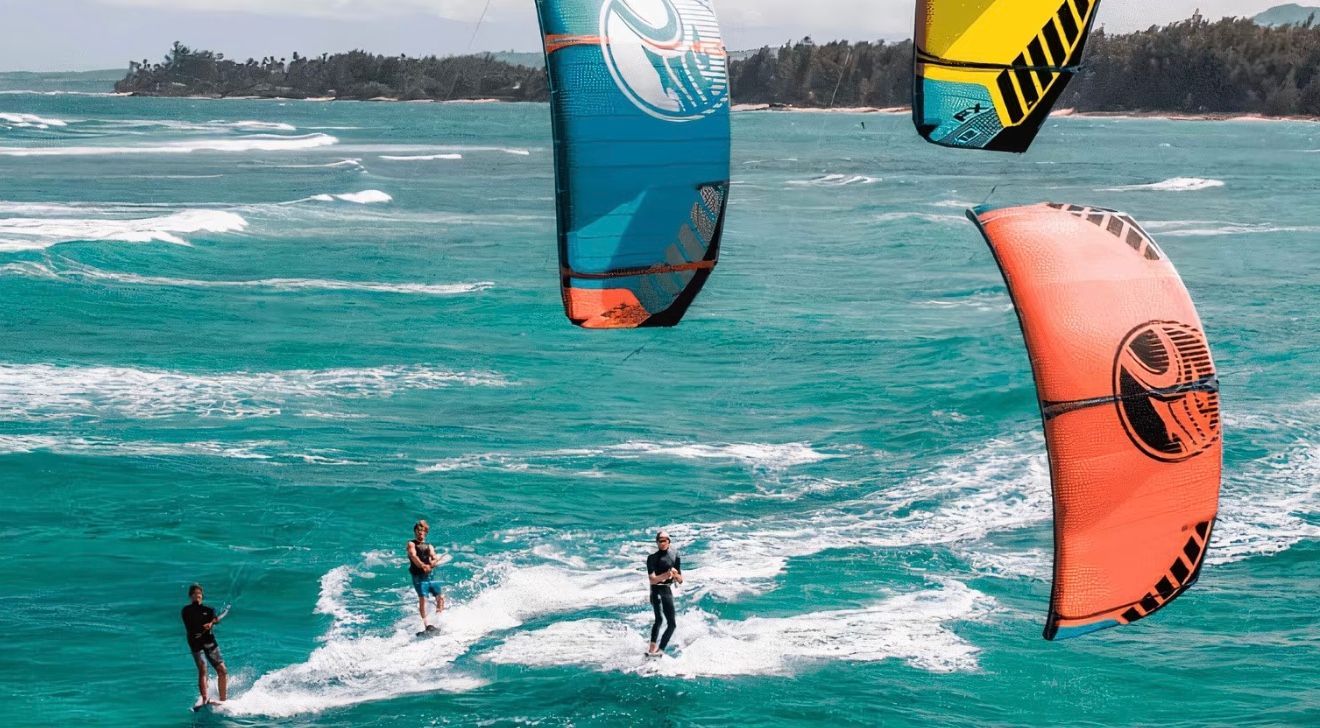 Kitesurfers on turquoise water with colorful kites against a tropical beach backdrop.