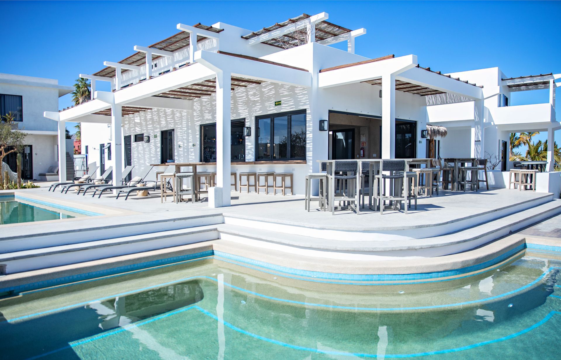 A modern white resort building with a patio, pool bar, and steps leading into a clear blue swimming pool.