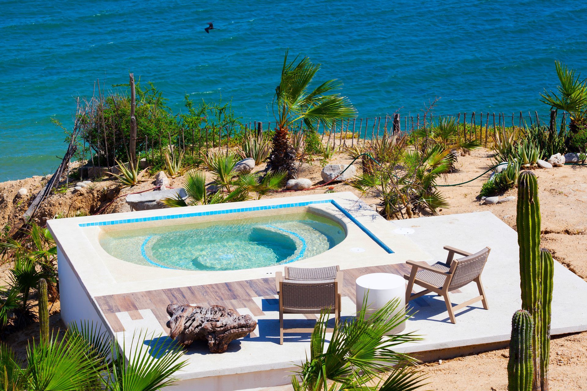 A small, round pool on a concrete patio overlooking the ocean, with two wooden chairs and desert plants nearby.