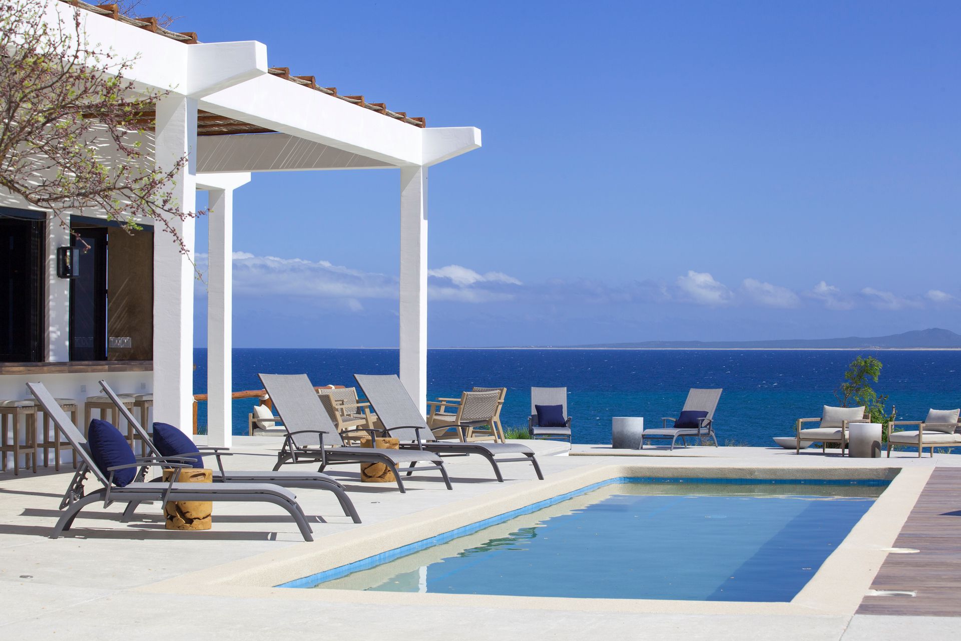 Pool deck with lounge chairs under a white pergola, overlooking a blue ocean and clear sky.