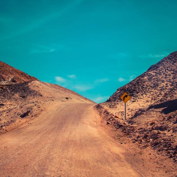 Dirt road winding between two hills, blue sky, yellow curve sign.