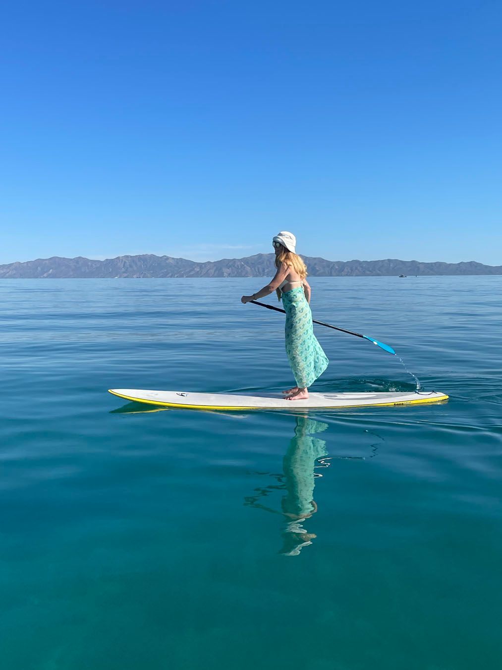 Woman paddleboards on turquoise water, mountains in the distance, under a clear blue sky.