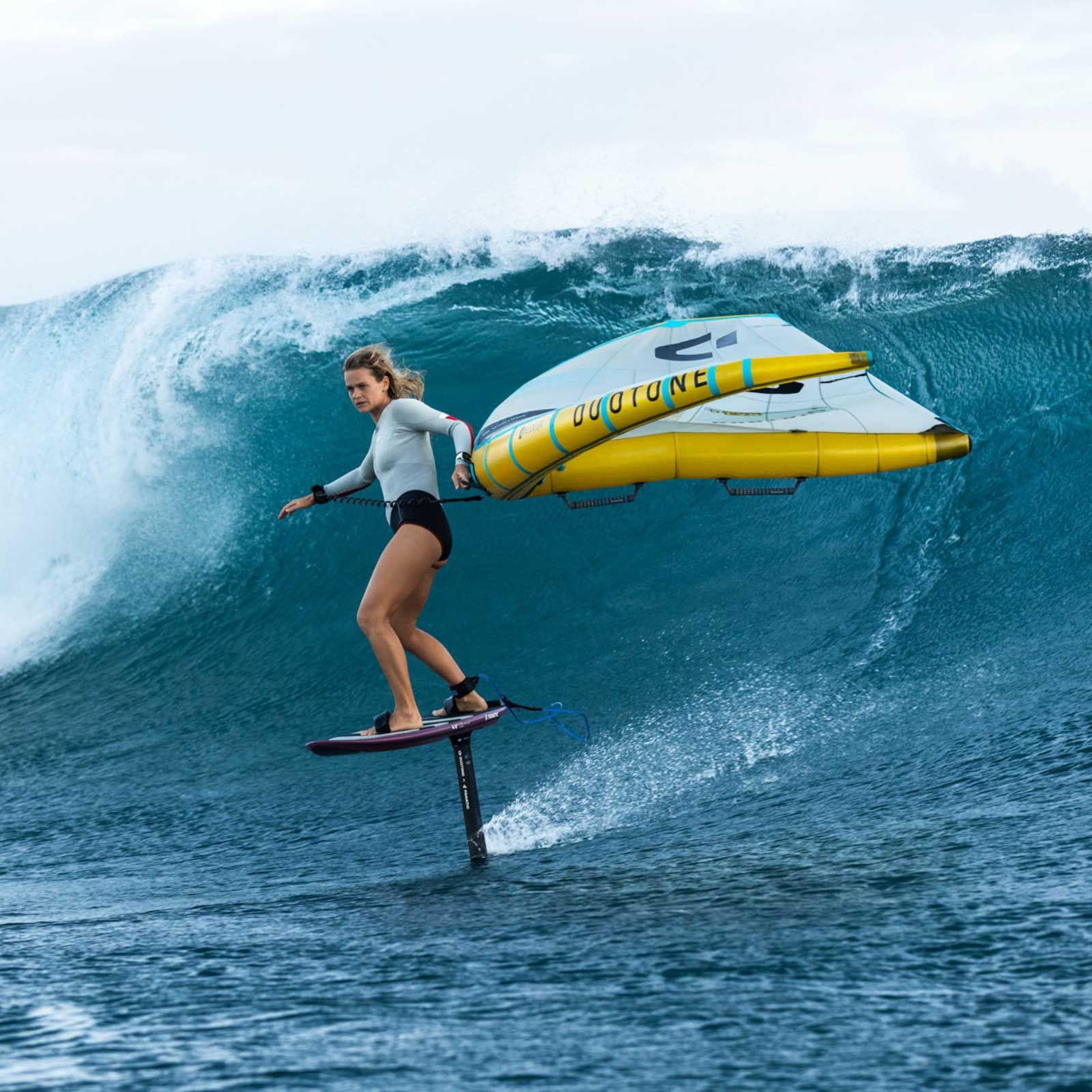 Woman wing foiling on a foil board, using a yellow wing in the ocean, with a wave in the background.