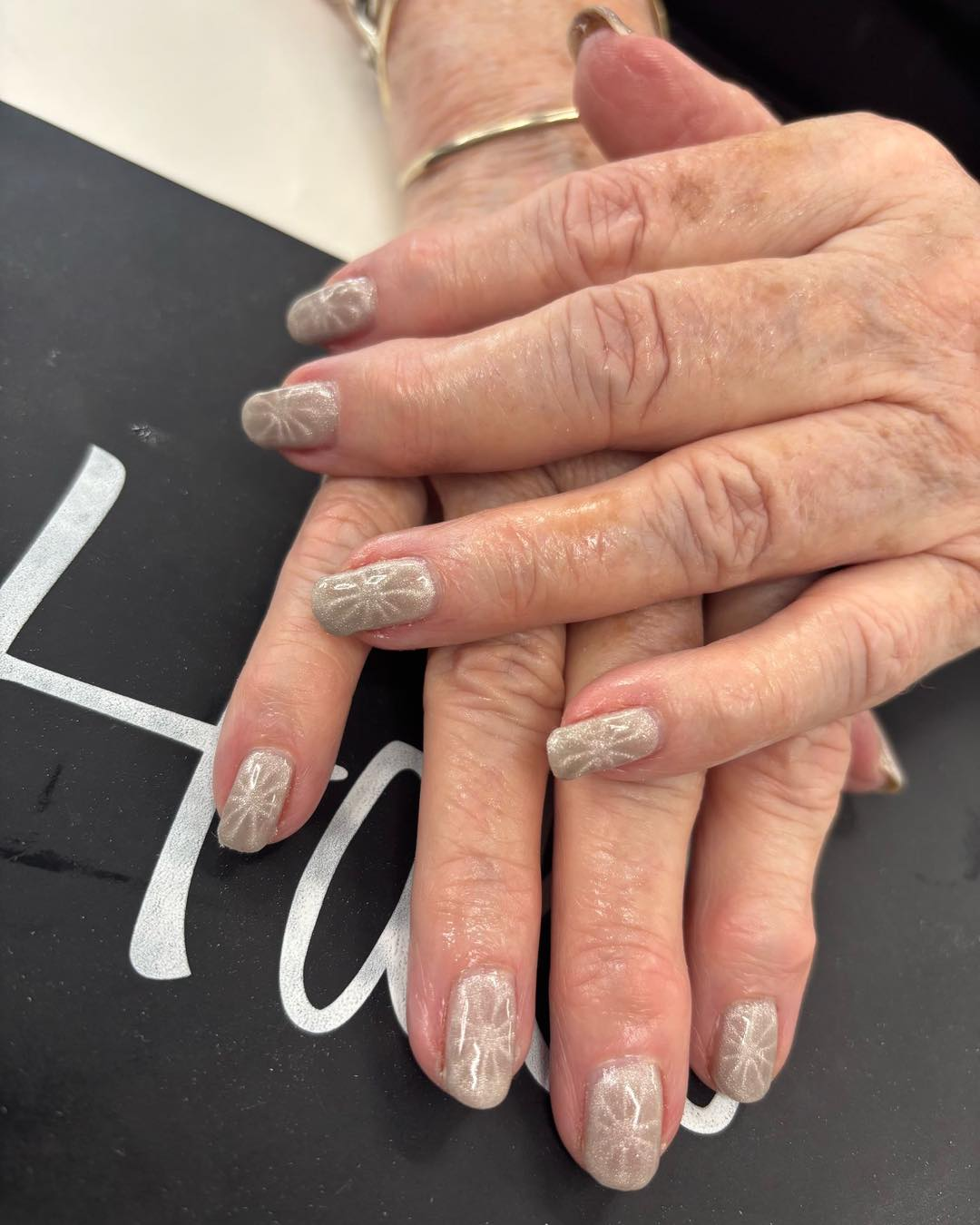 A pair of hands resting on a dark surface, showcasing manicured fingernails with a light-colored, patterned design.