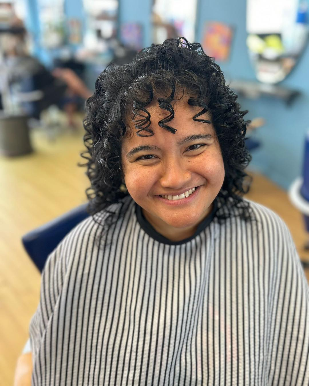 A person with dark, curly hair and bangs smiles in a hair salon, wearing a striped cape.