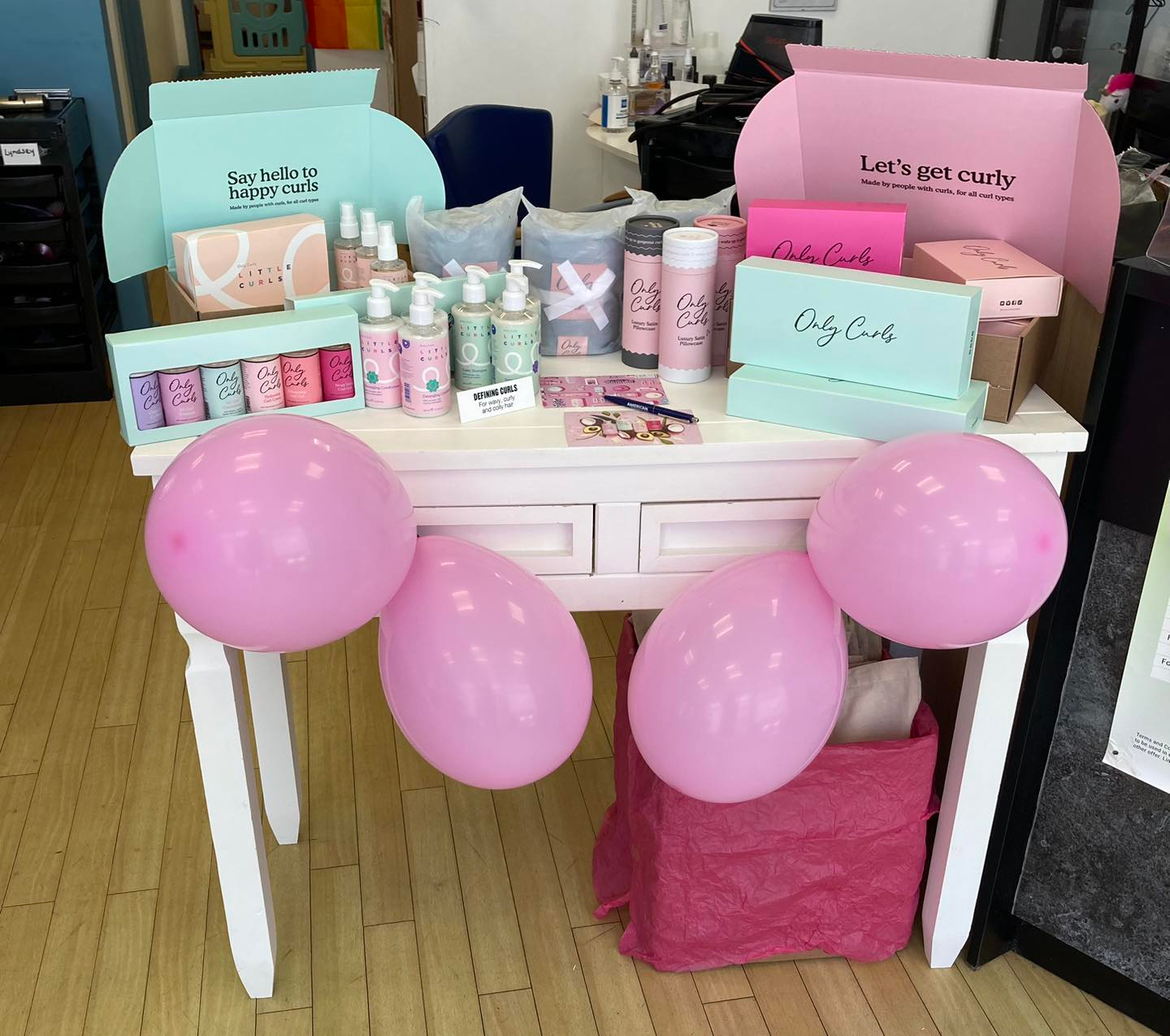 A white desk displays various pastel skincare products and boxes, decorated with pink balloons and a matching pink bin.