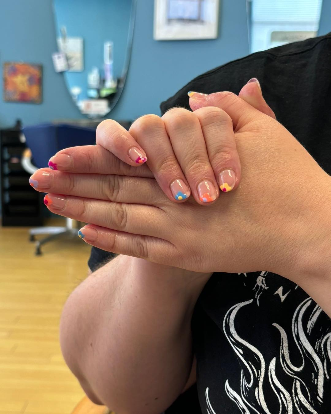 Hands are clasped to display short, natural nails with a clear base and colorful flower art on each tip in a salon setting.