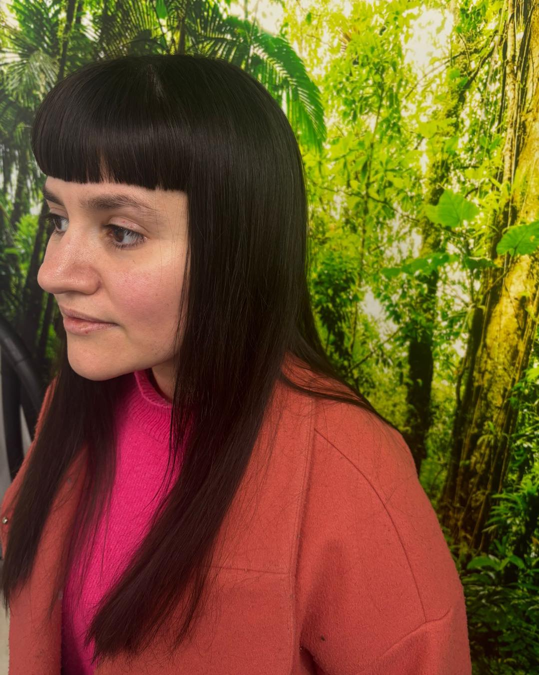 A person with dark, shoulder-length hair and straight-cut bangs wears a bright pink top and a rust-colored jacket.
