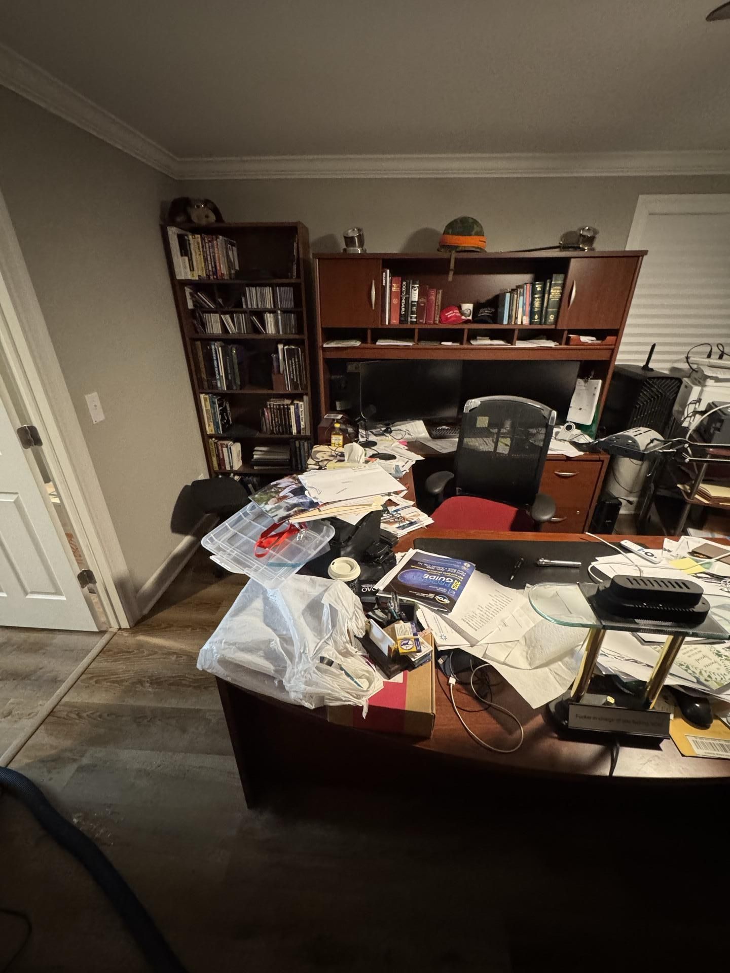 A cluttered home office with a wooden desk, hutch, bookshelf, and chair, illuminated by warm overhead lighting.
