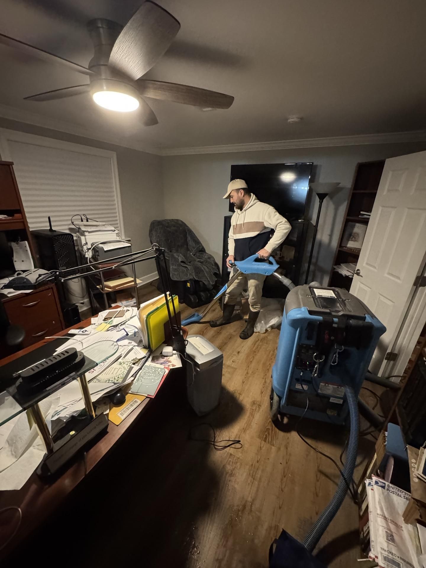 A worker uses a blue carpet cleaning machine to vacuum a room with hardwood floors, a desk, and a ceiling fan.