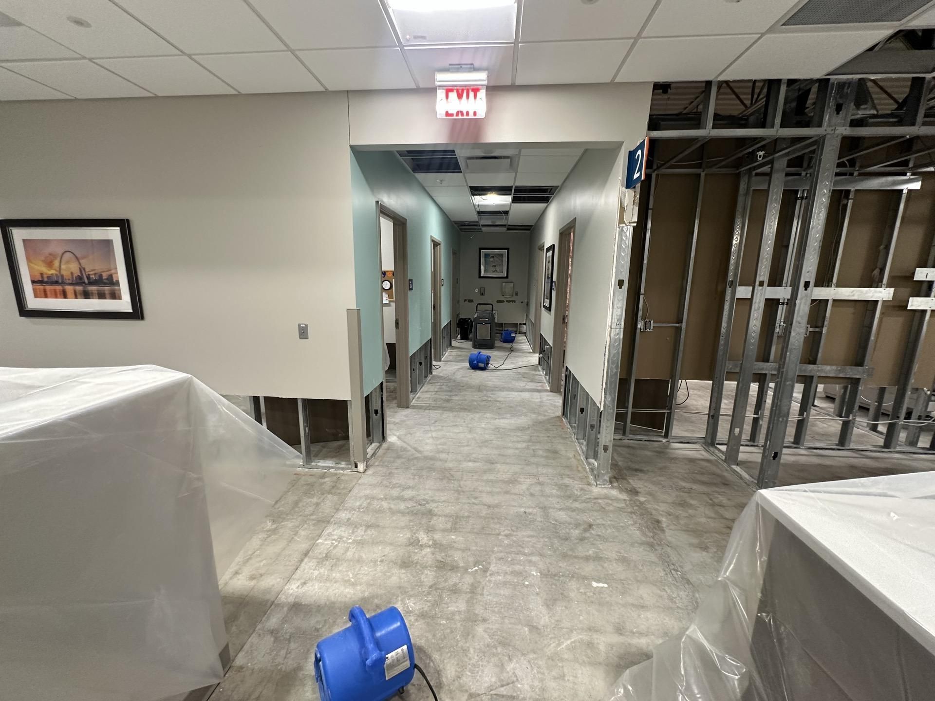 An office hallway under construction, featuring exposed metal studs, unfinished walls, and a blue floor blower fan.