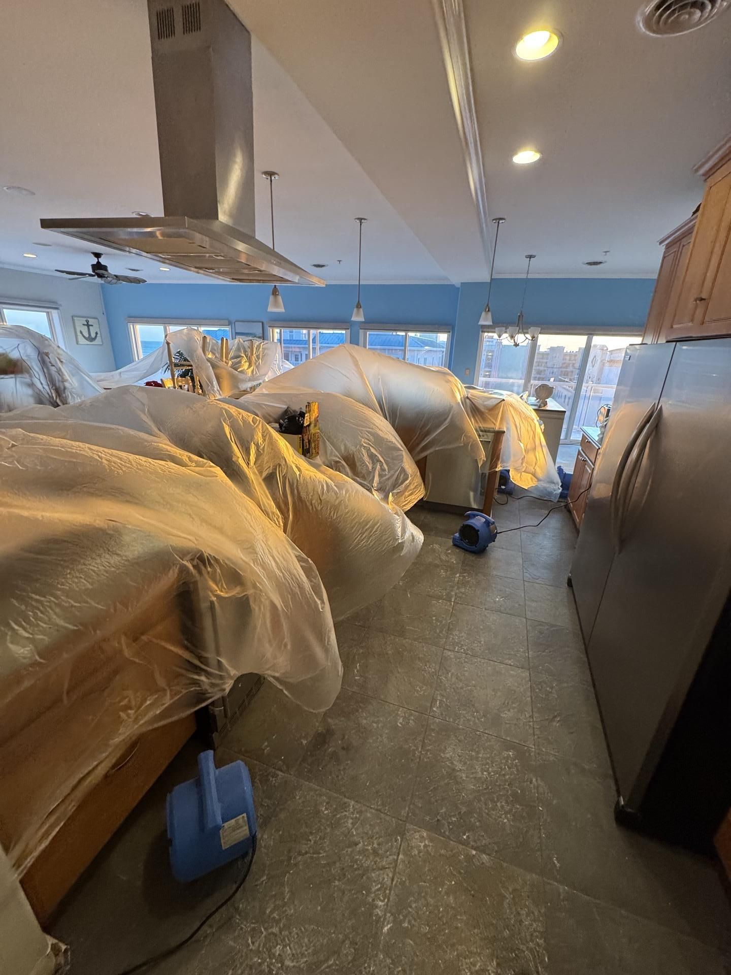 A kitchen undergoing renovation, with cabinets covered in plastic sheeting and industrial air movers on the tiled floor.