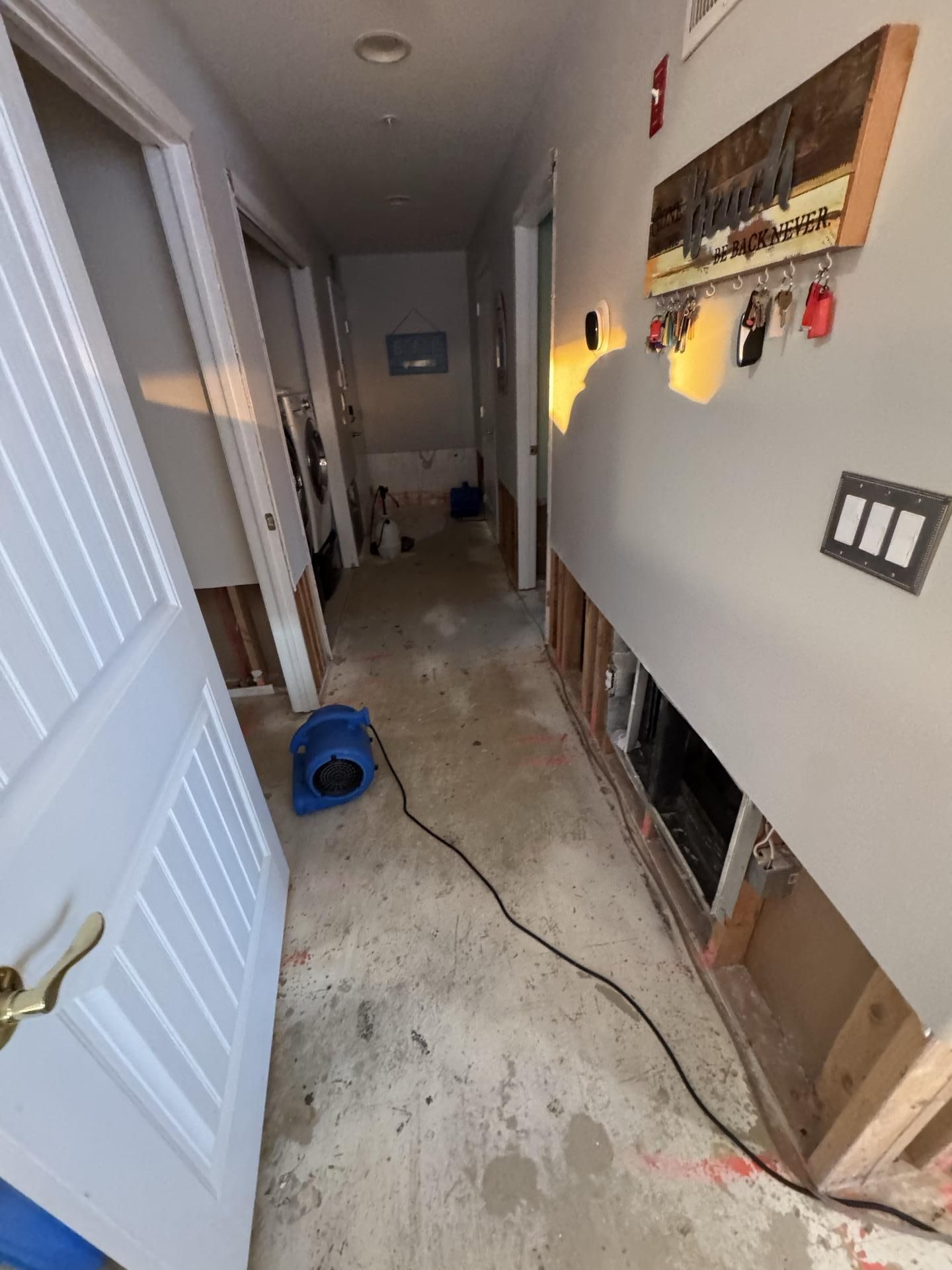 A hallway under renovation with removed baseboards, exposed wall studs, an air mover on the floor, and a key rack wall sign.