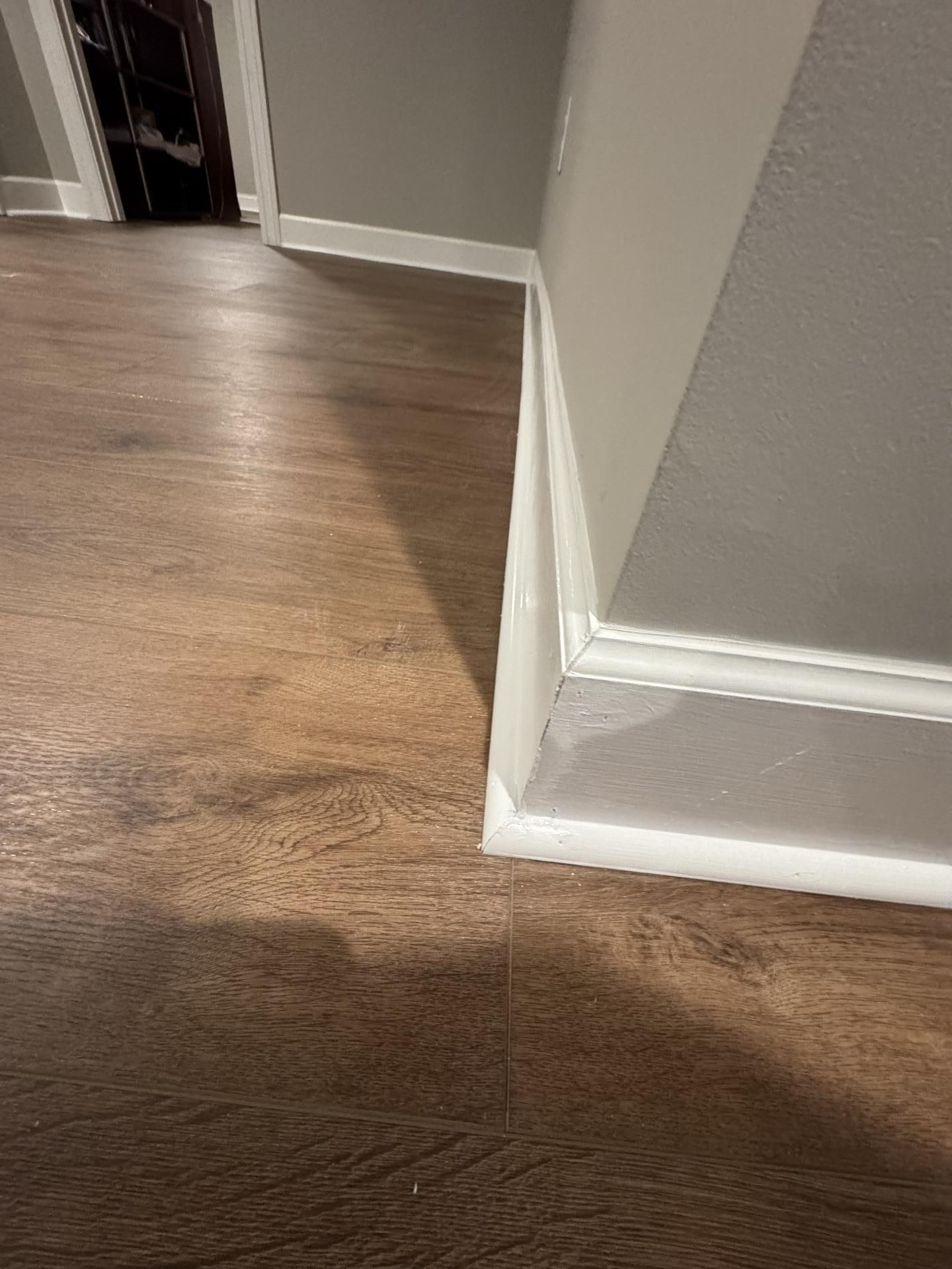 A corner of a room featuring wood-look flooring meeting white baseboard molding with a small gap along the floor.