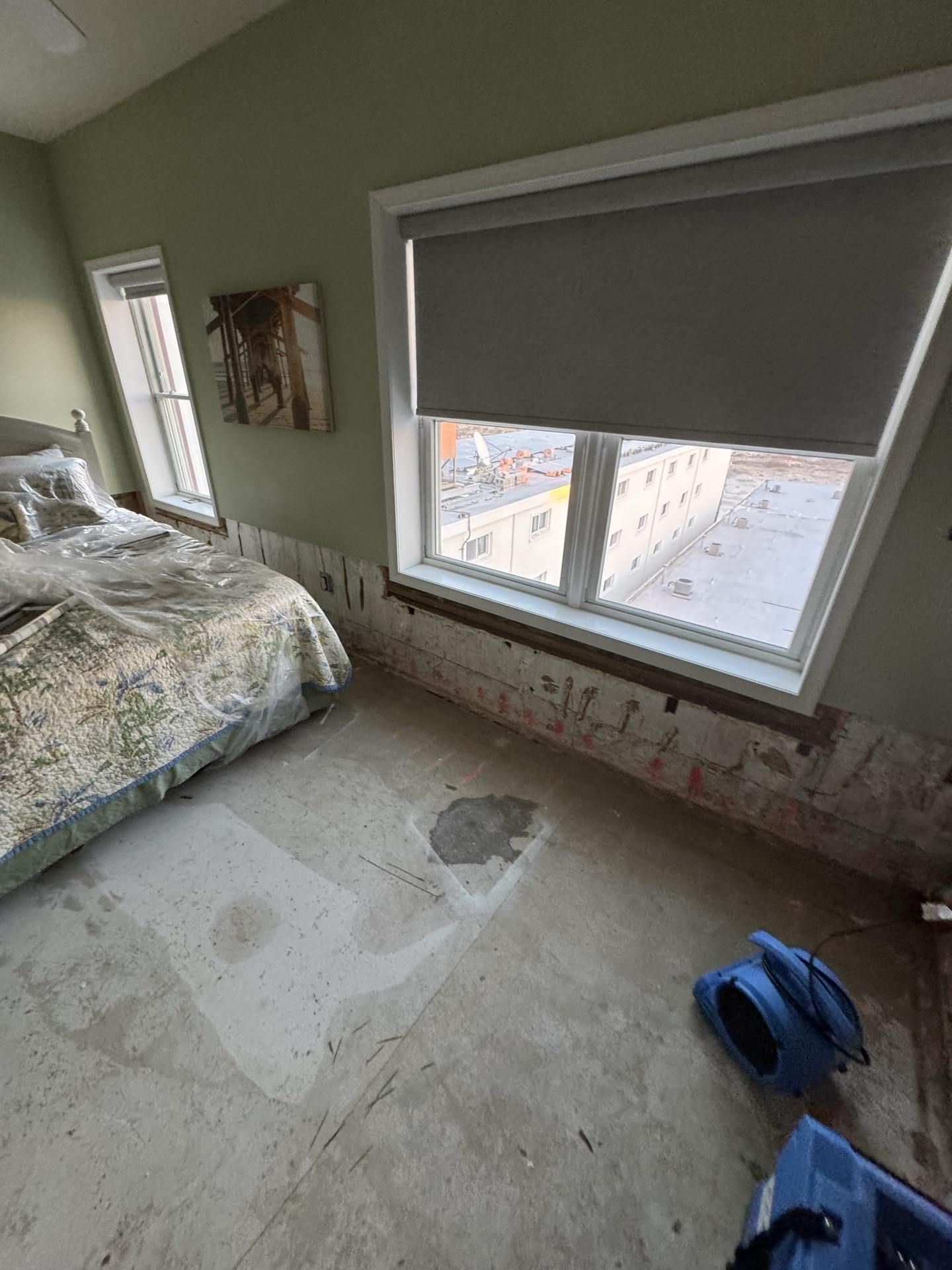 A bedroom under renovation with torn-out drywall, exposed subflooring, and an industrial air mover on the floor.