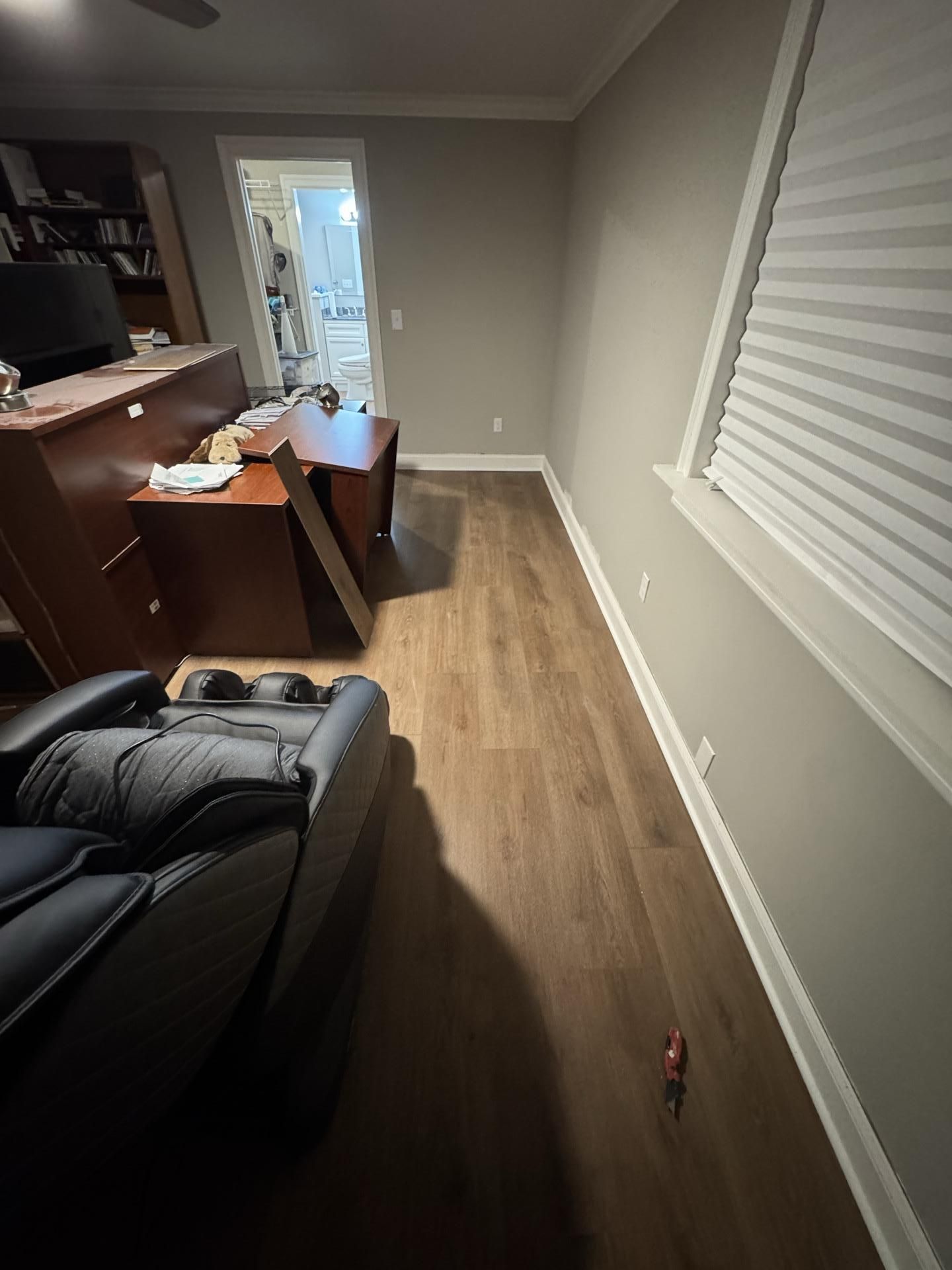 A dimly lit room featuring brown wood-look flooring, a large black recliner in the foreground, and a desk near a doorway.