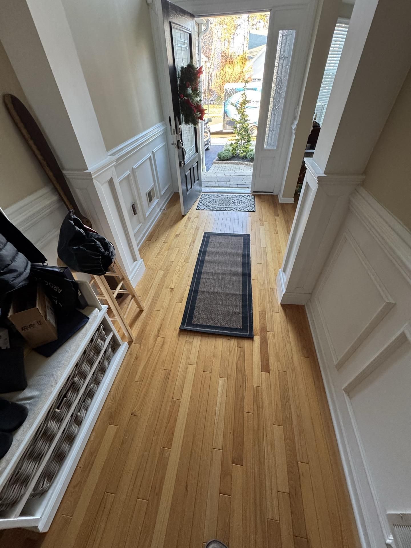 A bright entryway with light wood floors, white wainscoting, and a bench, leading to an open front door showing outdoors.