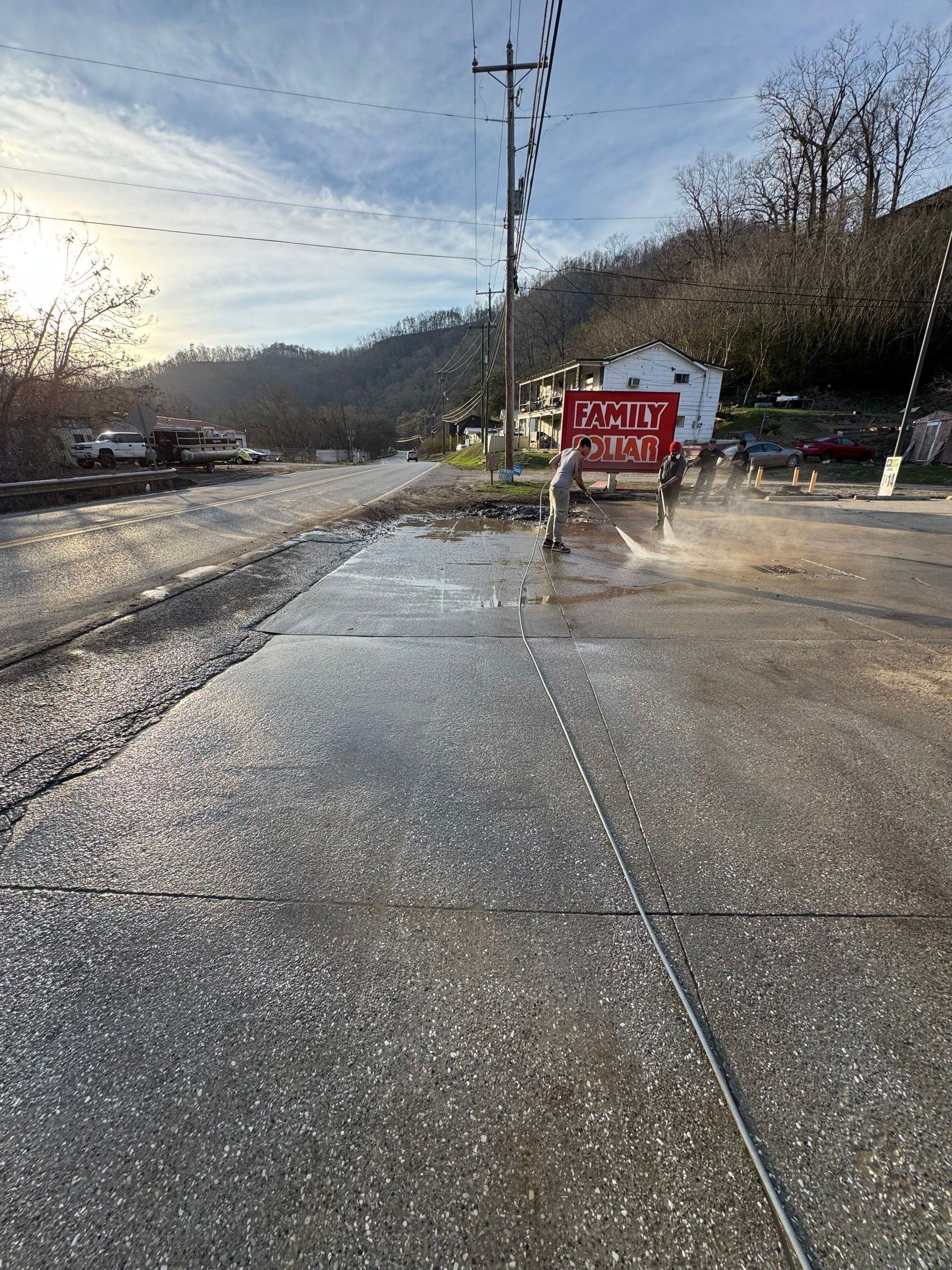 A person sprays water on a concrete lot covered in hail, with a red sign in the background against a hilly backdrop.