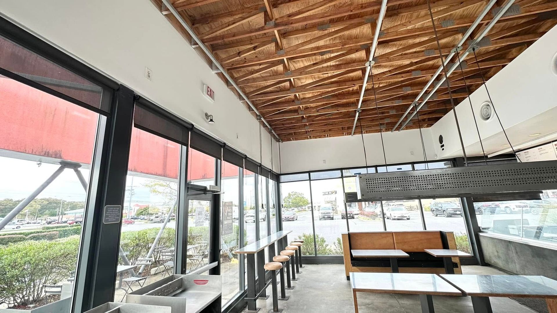 Modern restaurant interior with floor-to-ceiling windows, high-top seating, and a light-colored wooden exposed ceiling.