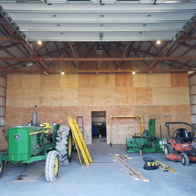 Farm equipment and tools in a metal workshop with plywood wall and open garage door