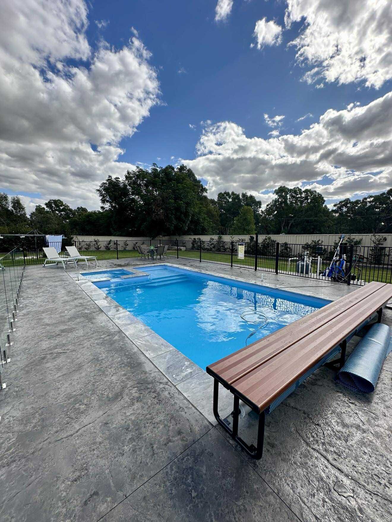 Swimming pool under a partly cloudy blue sky. Concrete patio with bench and trees in the background — Darlington Landscaping in Warren, NSW