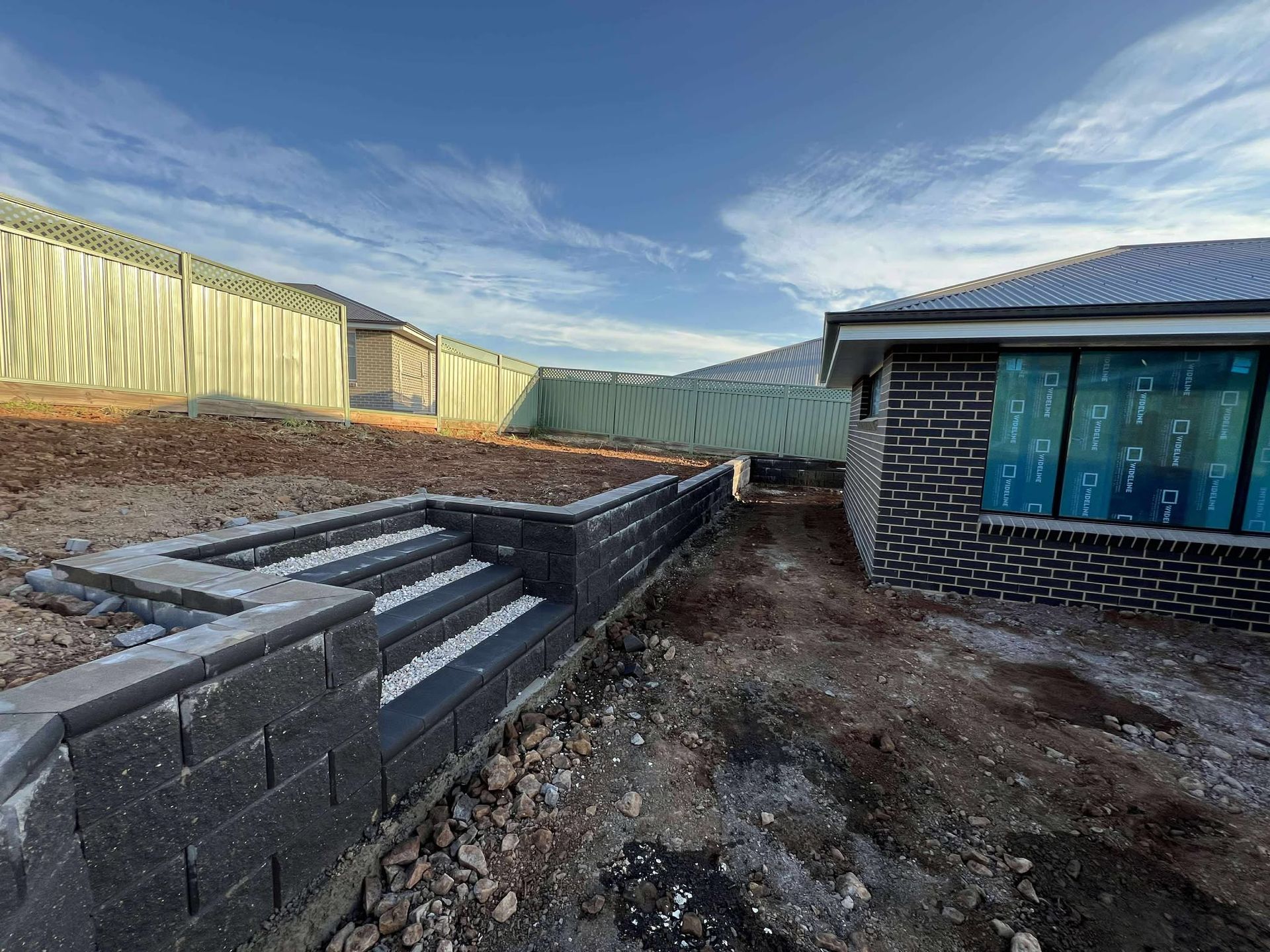 Backyard with retaining wall, steps, fence, and new brick house under construction — Darlington Landscaping in Dubbo, NSW