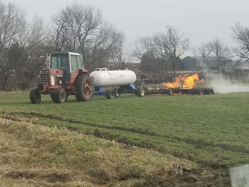 Tractor beside a white propane tank near a burning brush pile in a grassy field