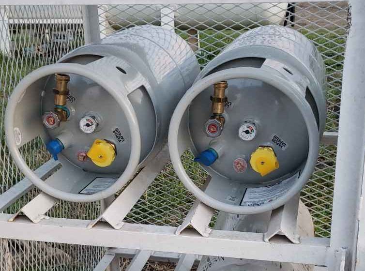 Two gray gas cylinders with valves, mounted on a white rack outdoors against a chain-link fence
