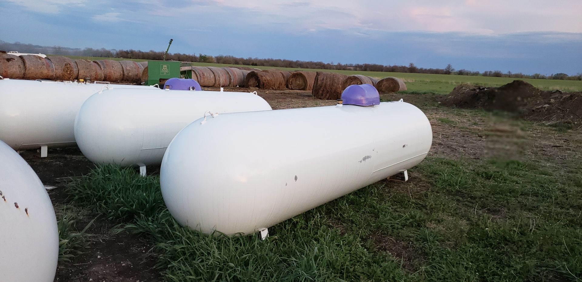 White propane tanks in a grassy field at sunset, with purple covers on some valves