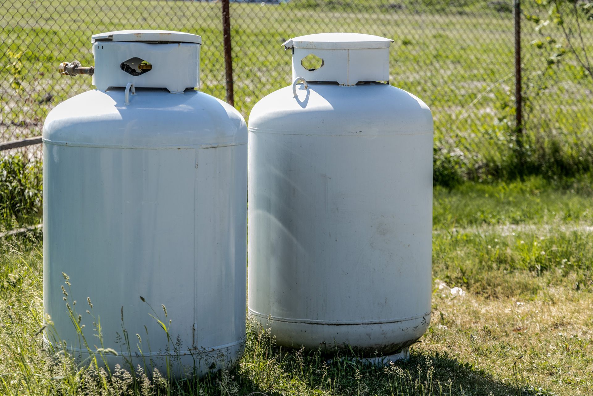 Two white propane tanks standing on grass beside a chain-link fence